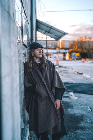 Moody urban portrait of someone in a dark coat and cap against an industrial building at sunset.