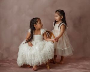 Two young children in elegant dresses share a sweet moment during a portrait session against a brown backdrop.