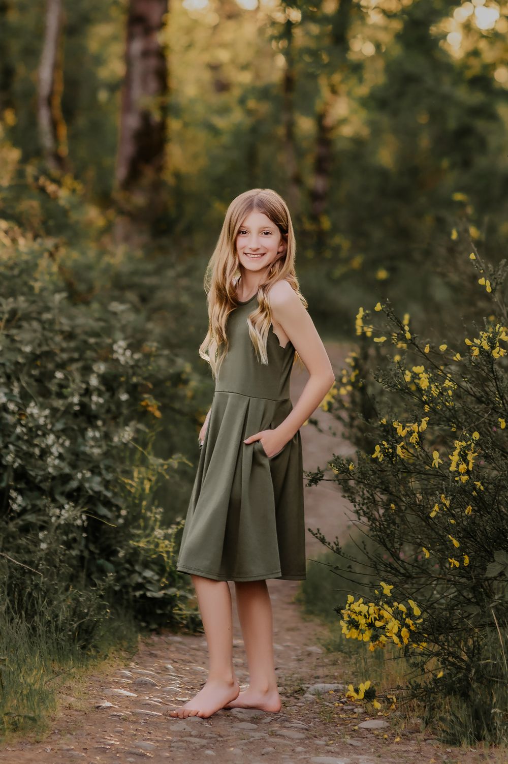 A barefoot young person in an olive green dress poses on a nature path surrounded by yellow wildflowers.