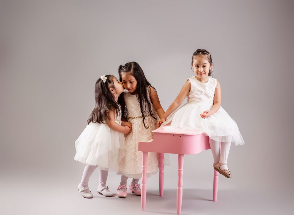 Three children in white dresses gather around a pink piano against a gray studio backdrop.