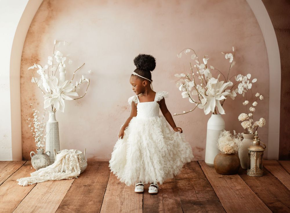 Child in white ball gown poses in arched alcove with dried floral arrangements.