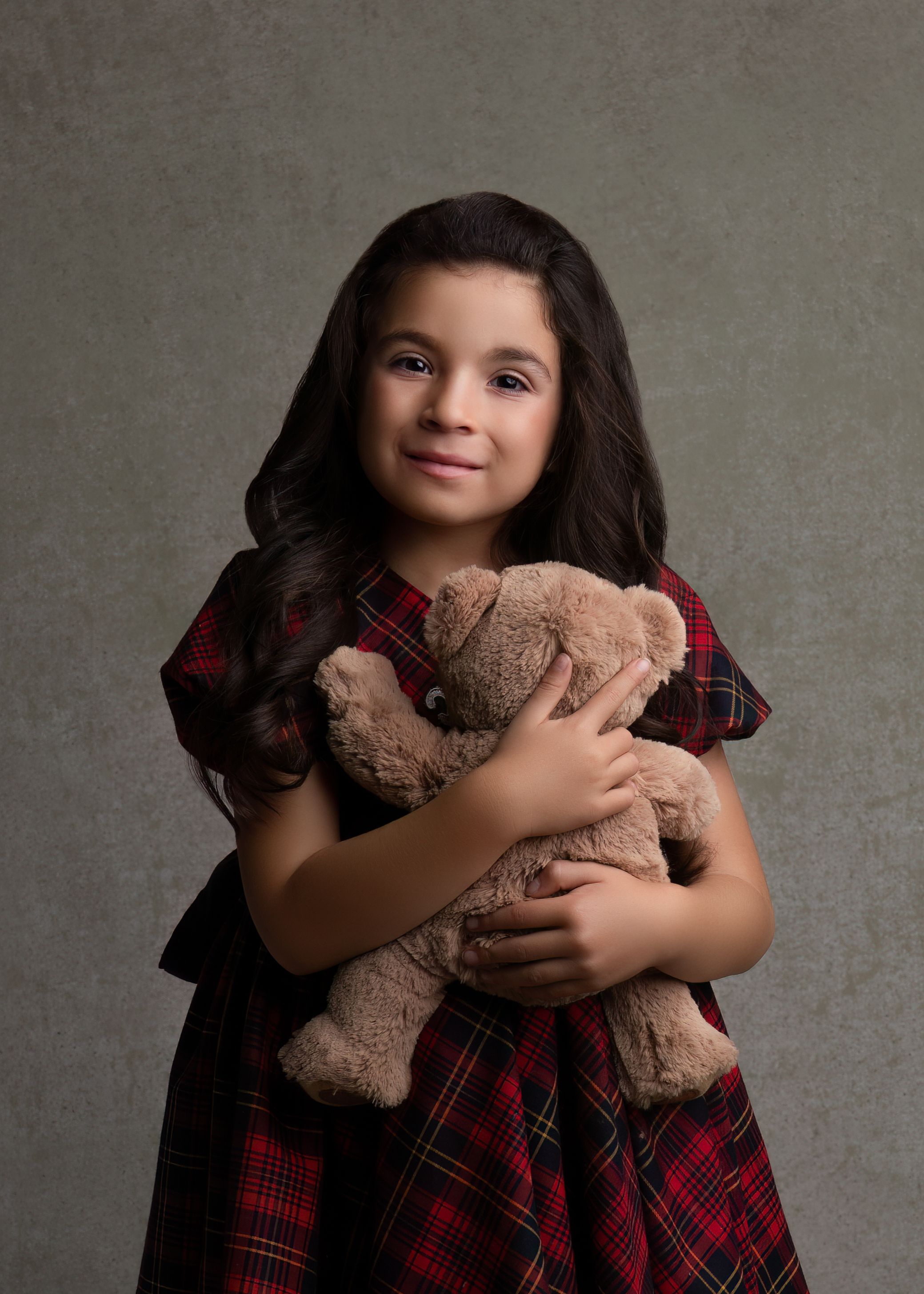 Studio portrait of young girl in plaid dress hugging teddy bear against gray background.