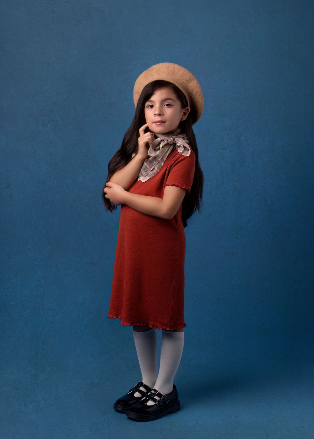 Portrait of girl in red dress and beret with floral scarf against blue backdrop.