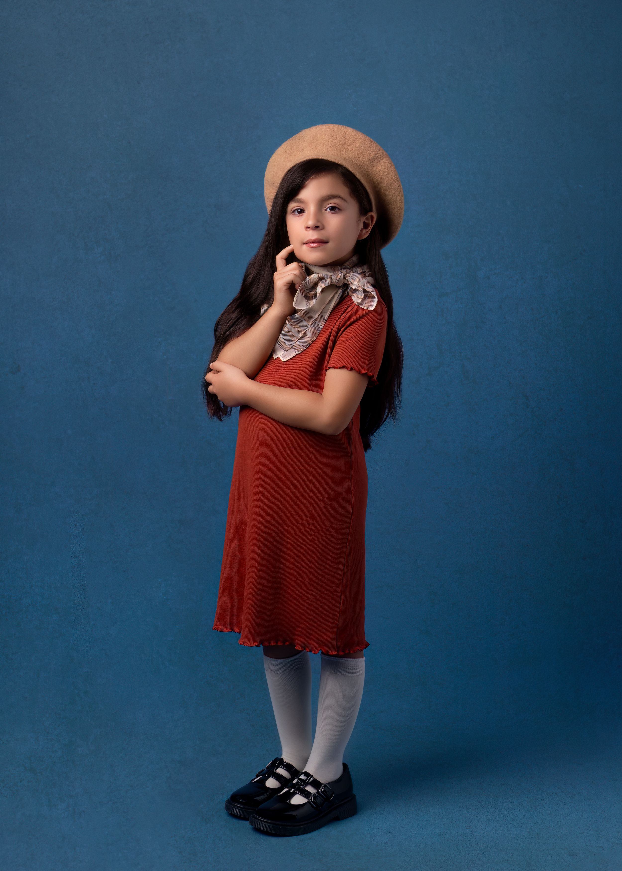 Portrait of girl in red dress and beret with floral scarf against blue backdrop.
