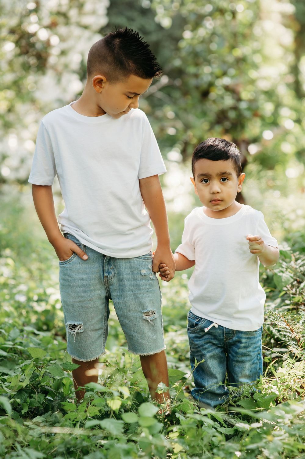 Two children in white t-shirts and denim hold hands while standing in lush green foliage.