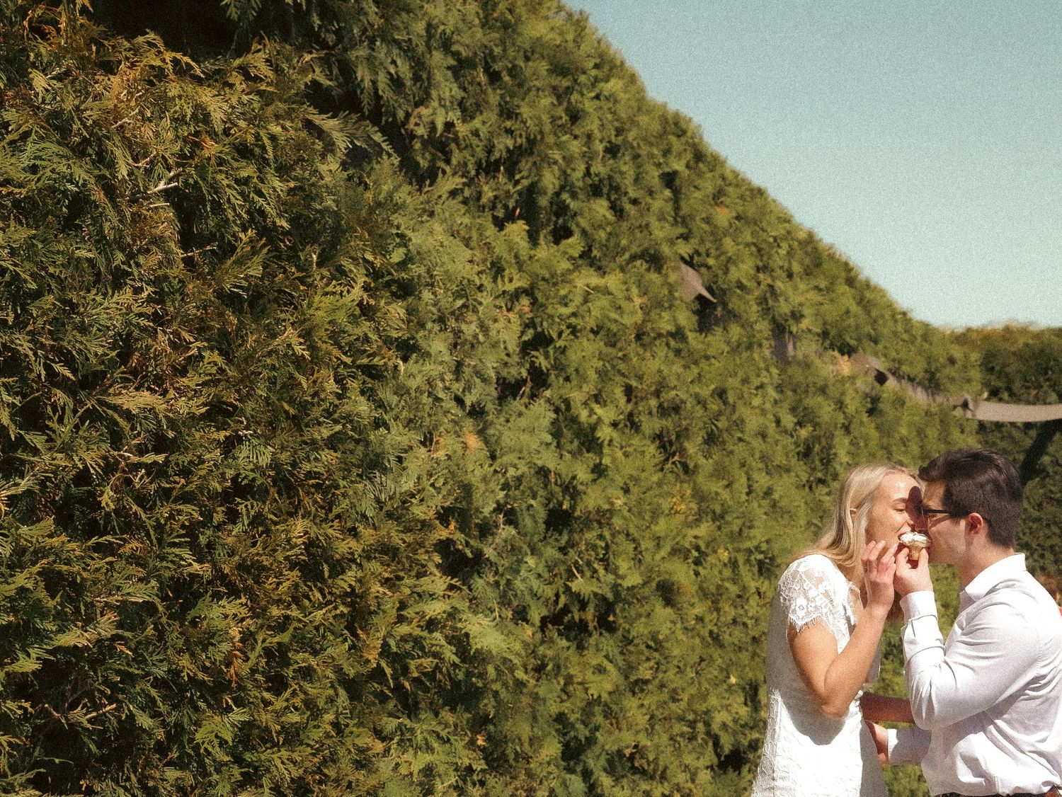 A couple shares a playful moment in front of a lush green hedge wall.