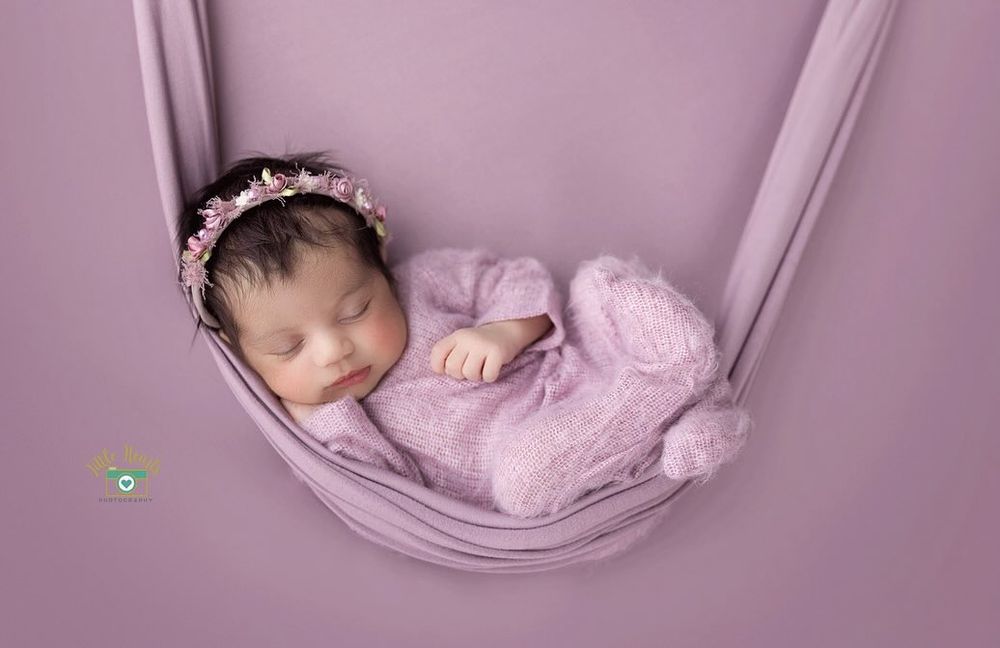 Sleeping newborn in pink wrap lying in fabric hammock against purple background.