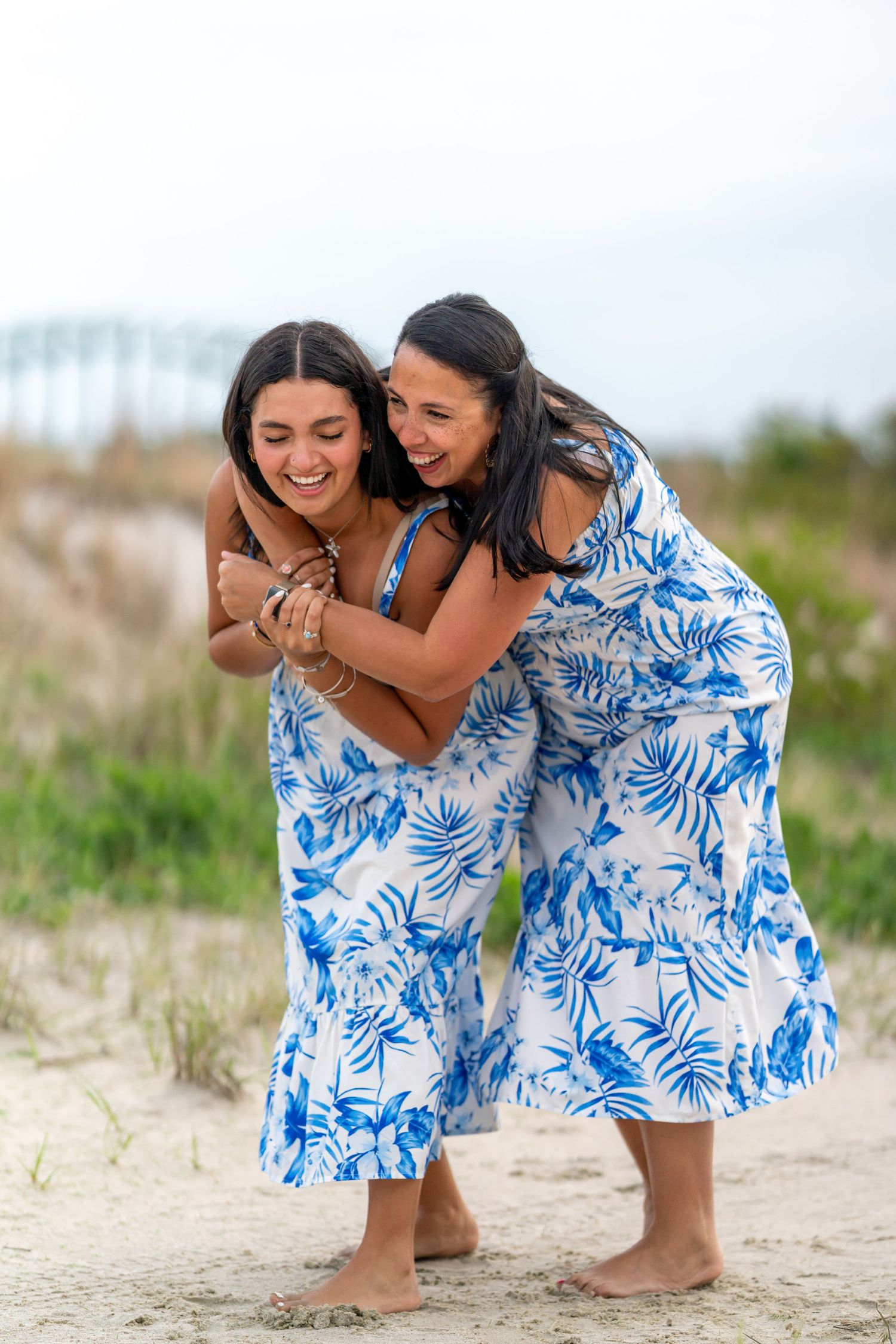 Two women in matching blue floral dresses laughing together on a beach.