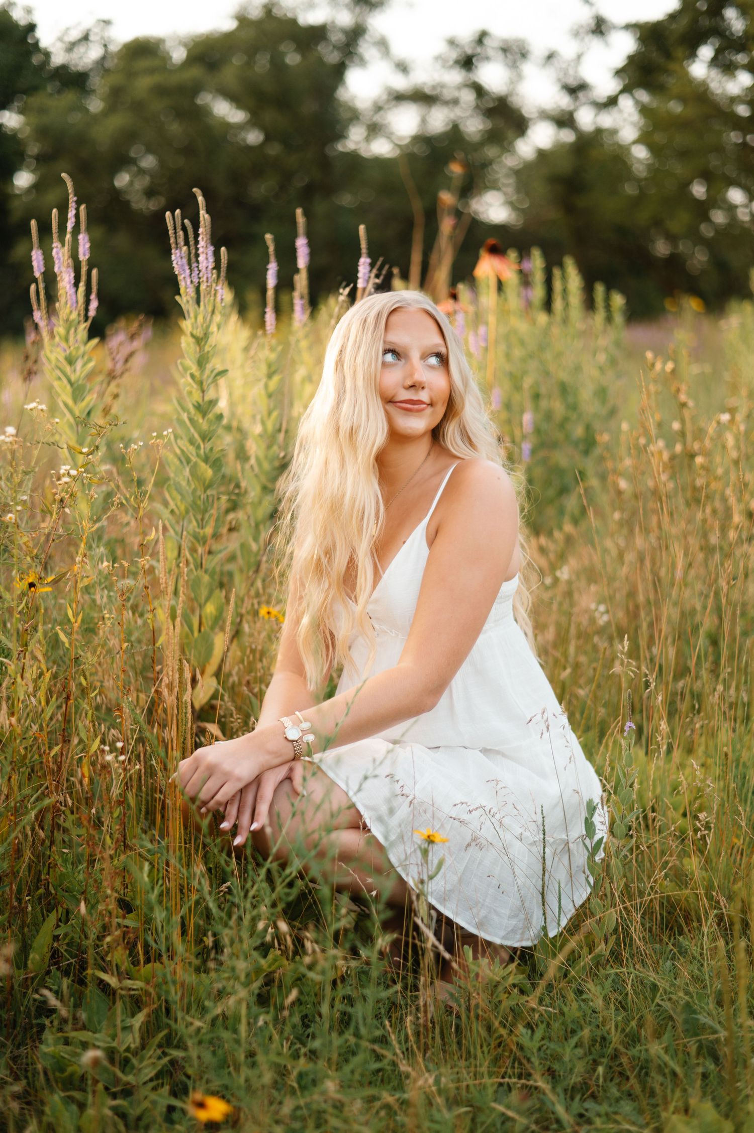 Blonde woman in white sundress crouching among wildflowers in golden meadow at sunset.