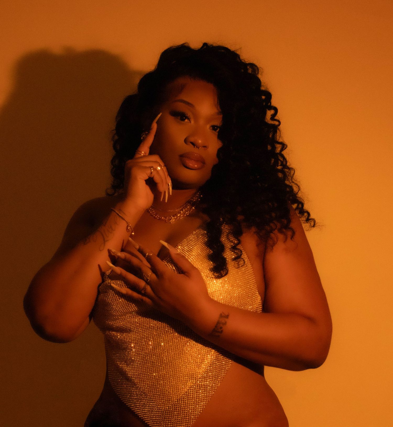 Woman in a gold sequin top poses against an orange-lit backdrop with long curly hair and dramatic lighting.