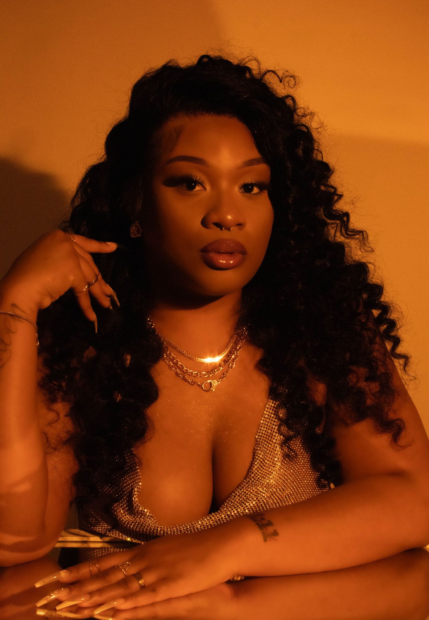 A portrait in warm amber lighting featuring glamorous curly hair, jewelry and a sparkly top against an orange background.