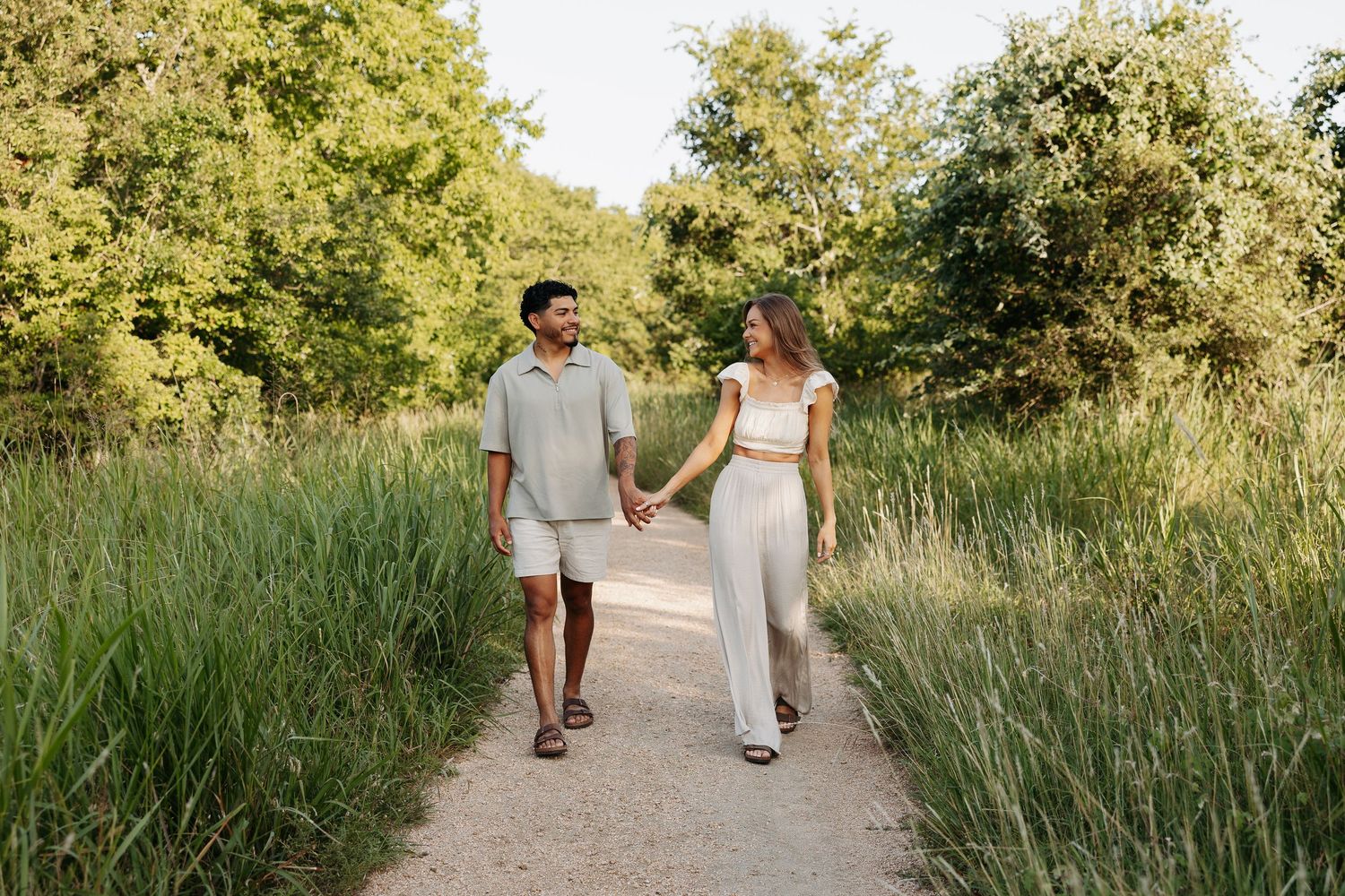 A couple walks hand in hand down a nature path surrounded by tall grass.