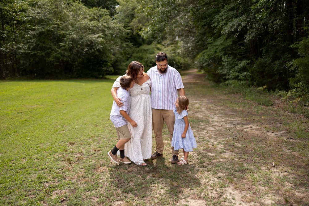 Family of four embracing on a scenic wooded path, parents hugging two young children in a lush green outdoor setting.