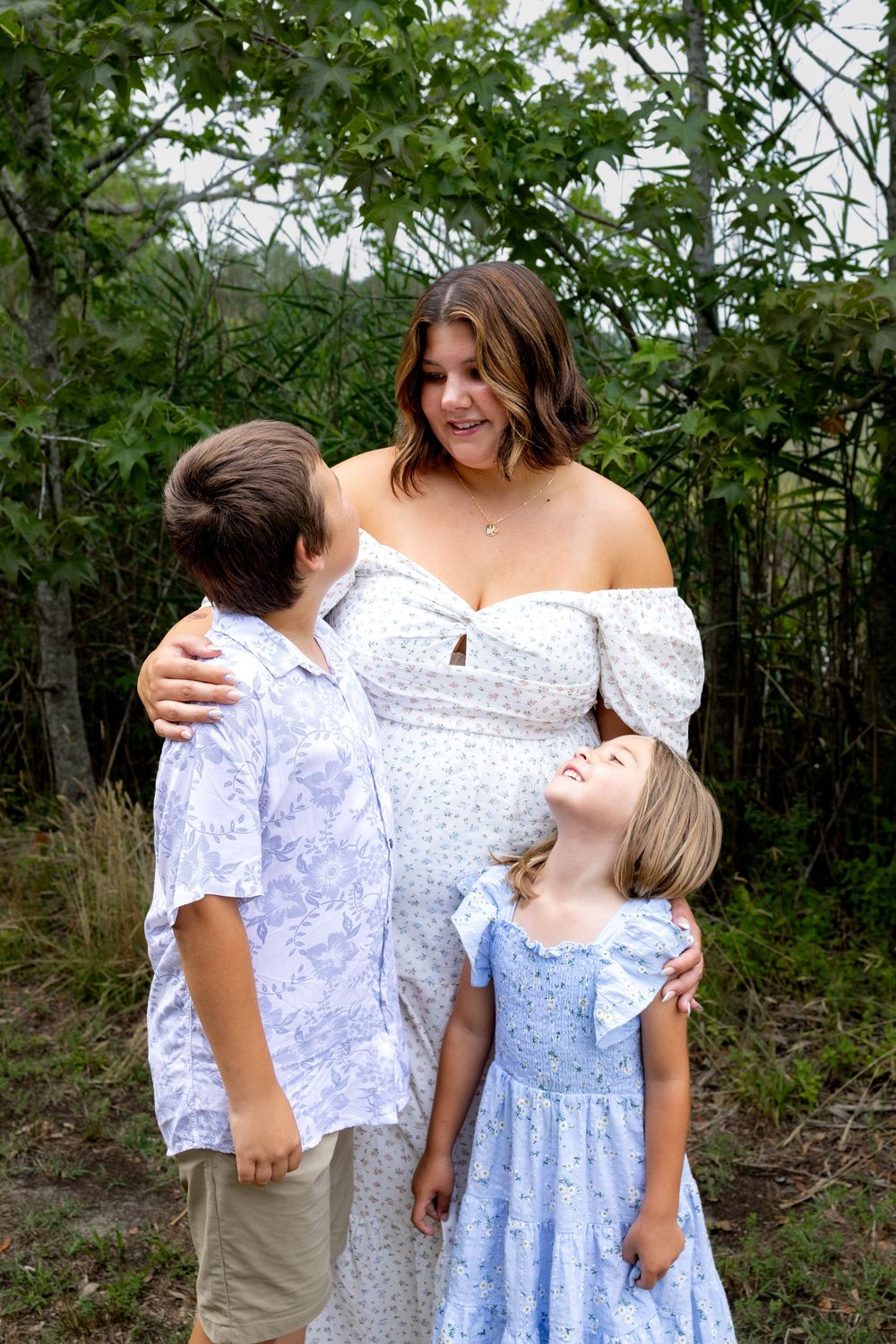 Pregnant woman in white floral dress smiles with two children outdoors among green trees.