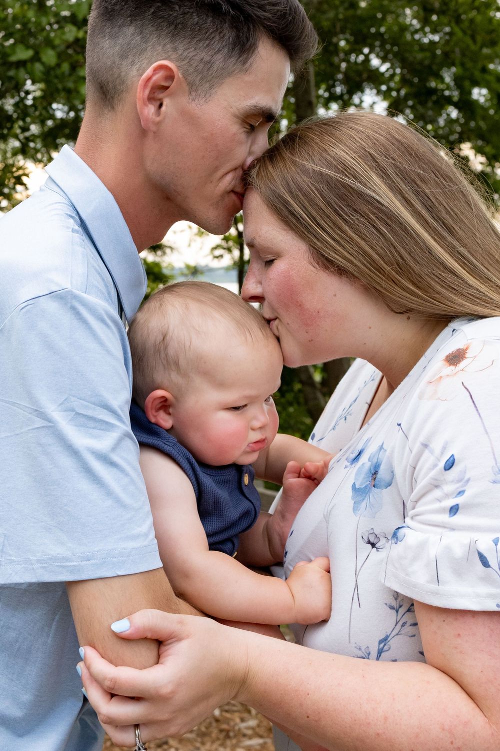 A couple kisses their baby boy outdoors, holding him tenderly in a warm family portrait.