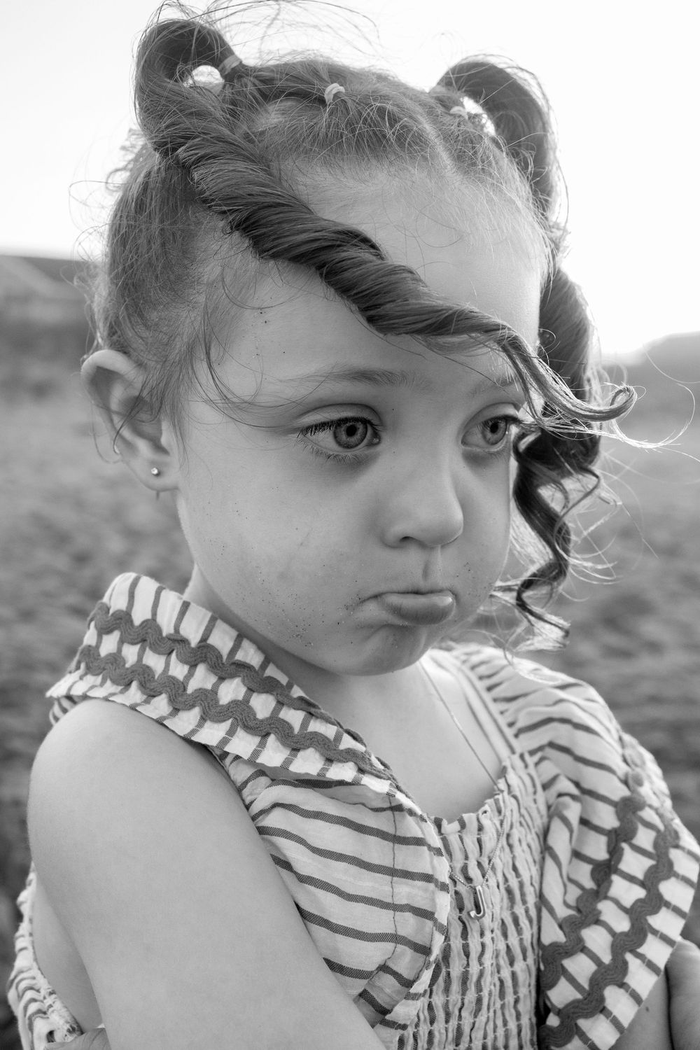 Black and white portrait of a young girl with pigtails and curly hair, wearing a striped dress, making a pouty face.