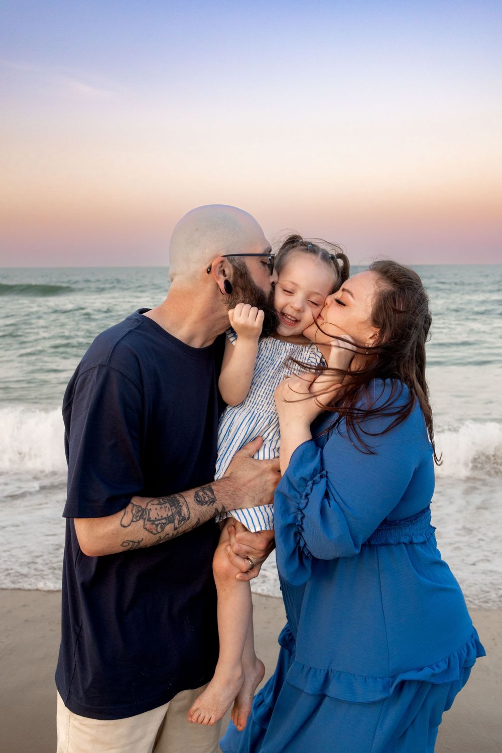 Happy family with tattooed dad and mom in blue dress kissing their laughing daughter on a beach at sunset.