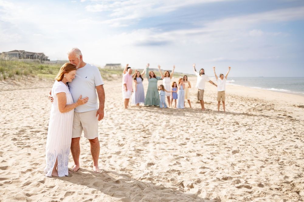 Couple poses on sandy beach while large family group celebrates behind them on a sunny day.