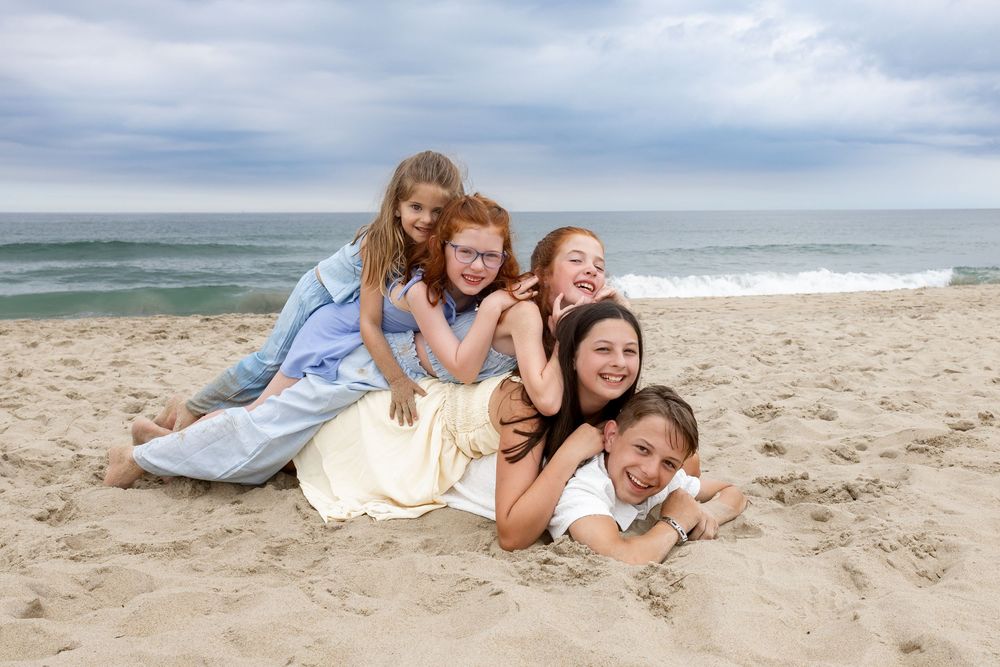 A joyful family of five piling on each other on a sandy beach with ocean waves in the background.