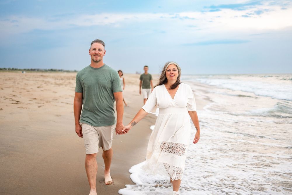 Couple holding hands walking on a sunny beach in casual attire, with two people visible in the background near the ocean.