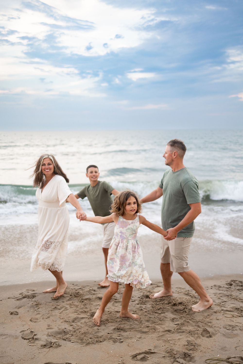 Happy family of four walking barefoot on a sandy beach, holding hands with ocean waves in the background.