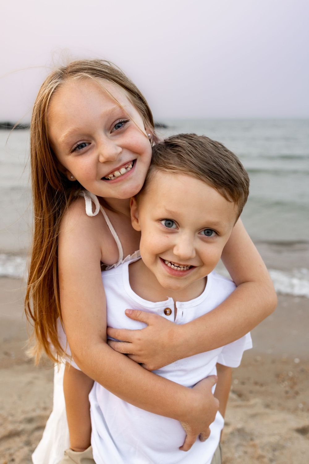 Two happy children hugging and smiling on a beach, girl with long hair embracing younger boy in white shirt.