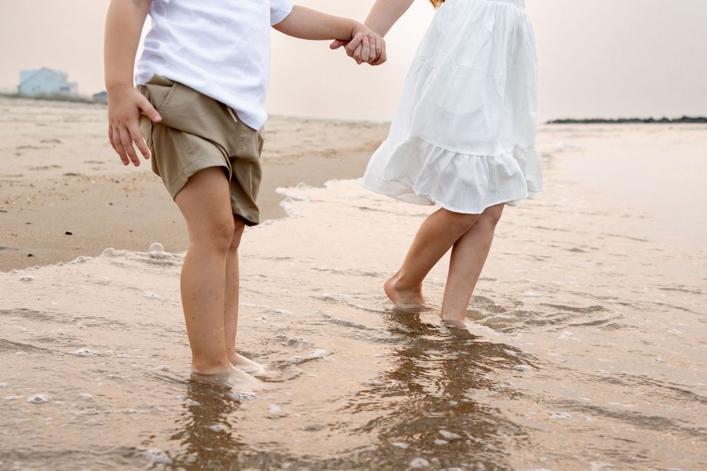 Two children holding hands while wading barefoot in ocean waves on a sandy beach.