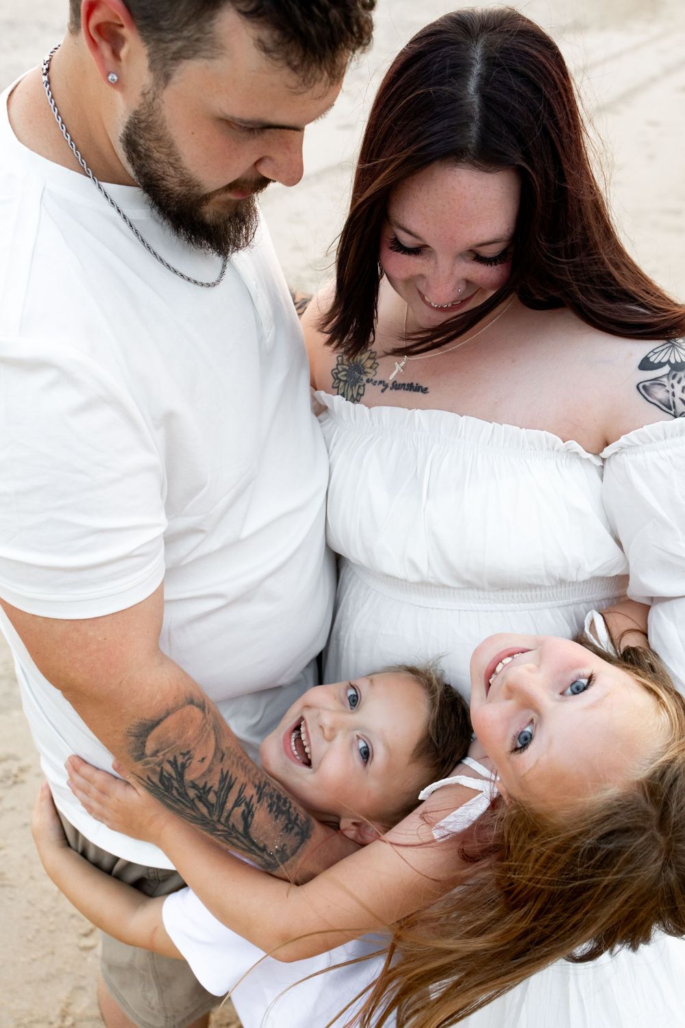 Family of four in white outfits sharing a joyful moment on a sandy beach, parents holding two laughing children.