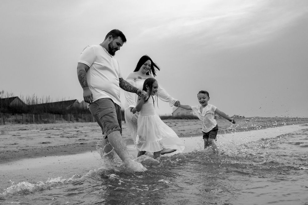 Black and white photo of a family of four joyfully splashing in ocean waves on a beach.
