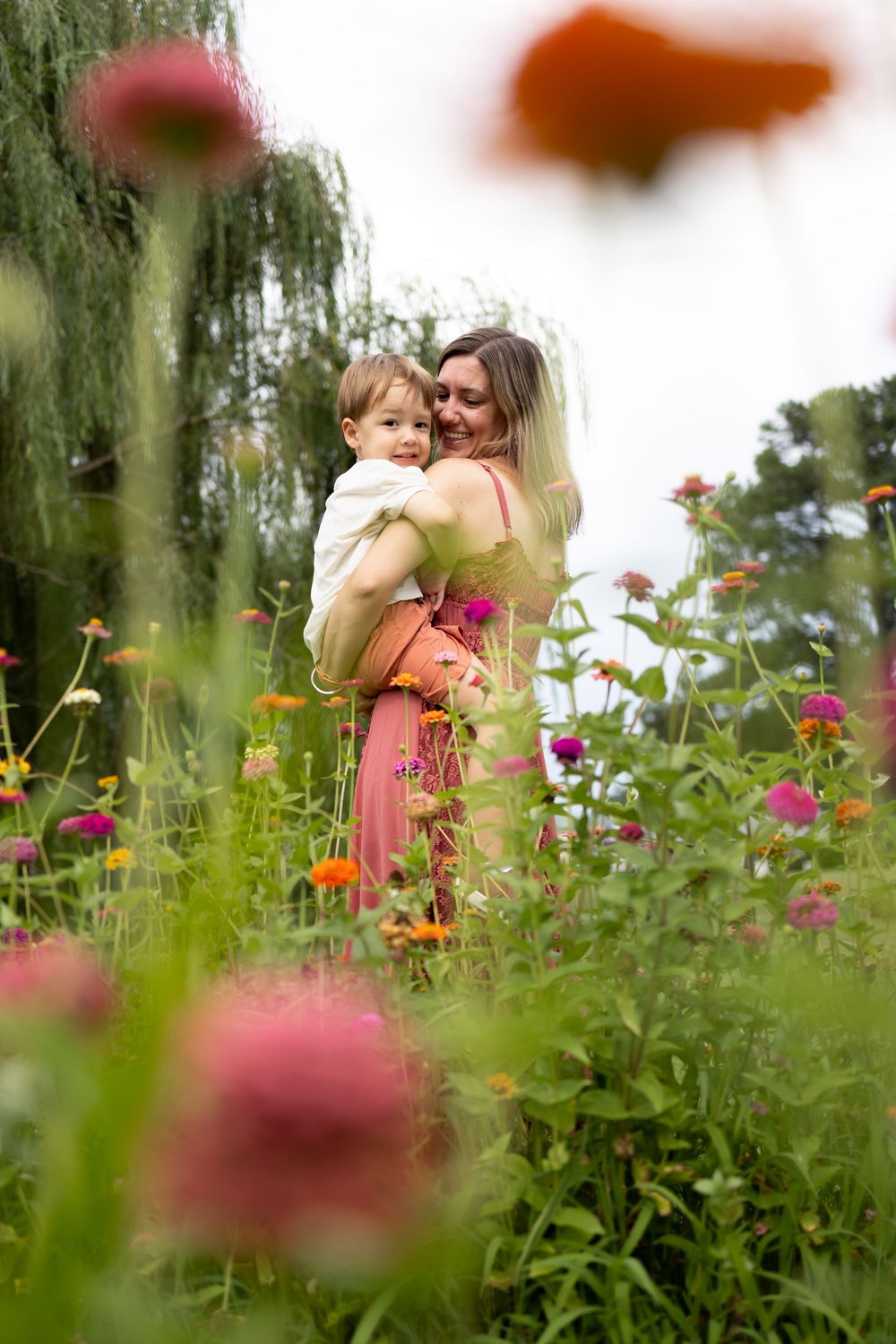 A mother in a pink dress holds her toddler in a vibrant flower garden with colorful blooms and lush green trees.