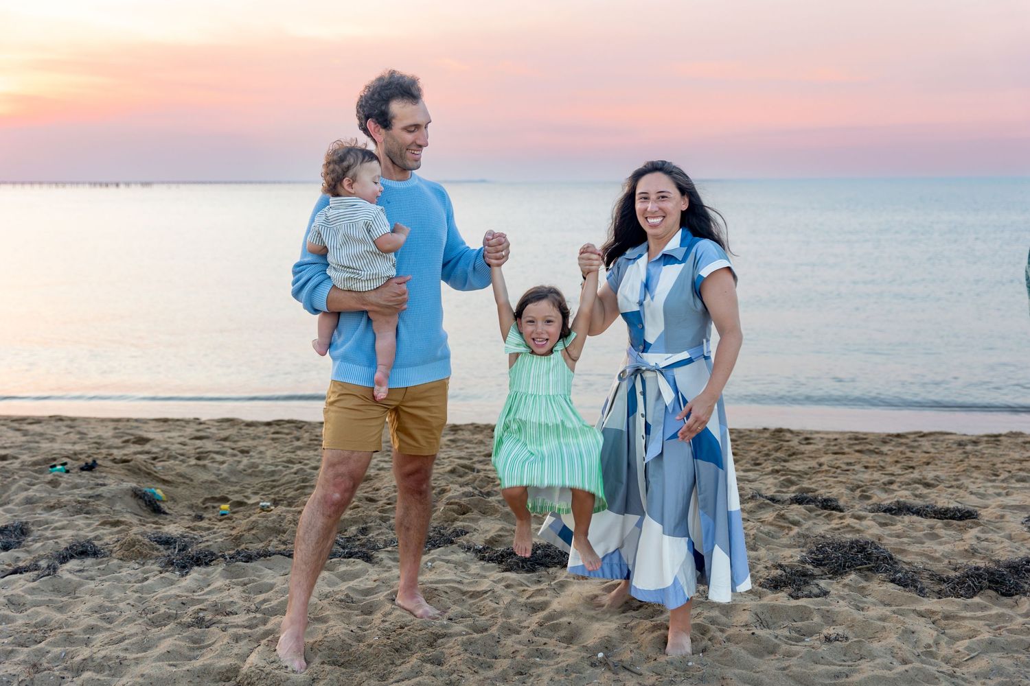 A family walks hand in hand along a sandy beach at sunset while waves crash against the shore.