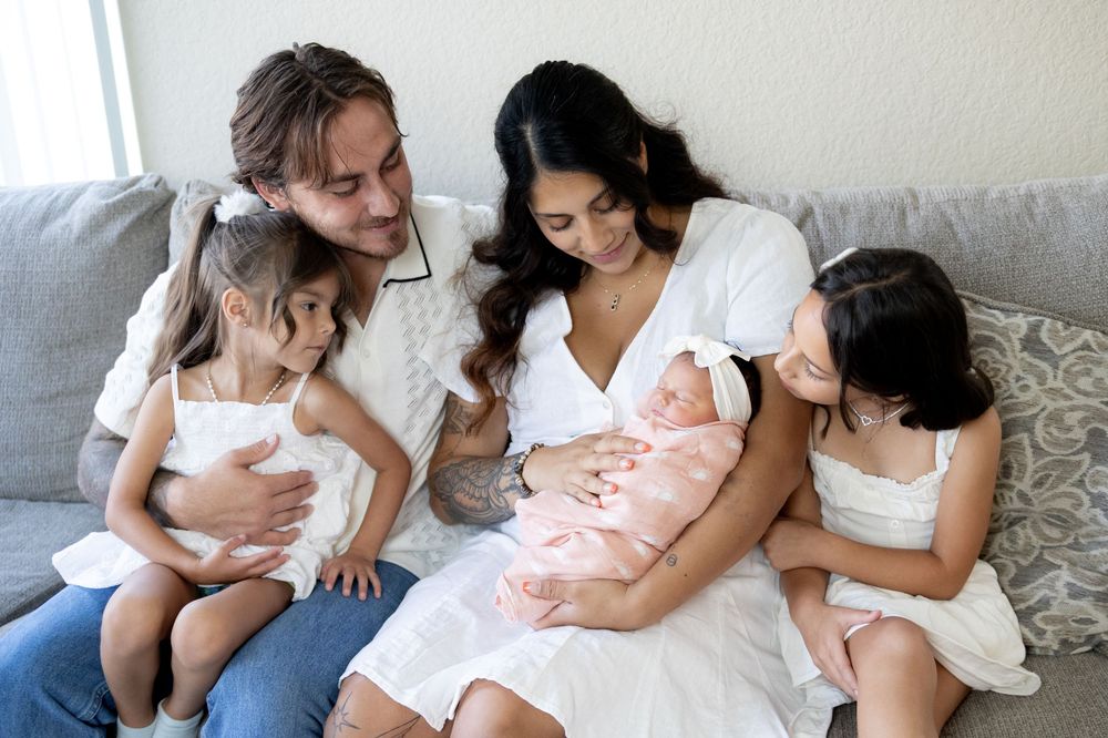 A tender moment shared on a gray couch, as loved ones gather close to coo over a newborn baby in pink.