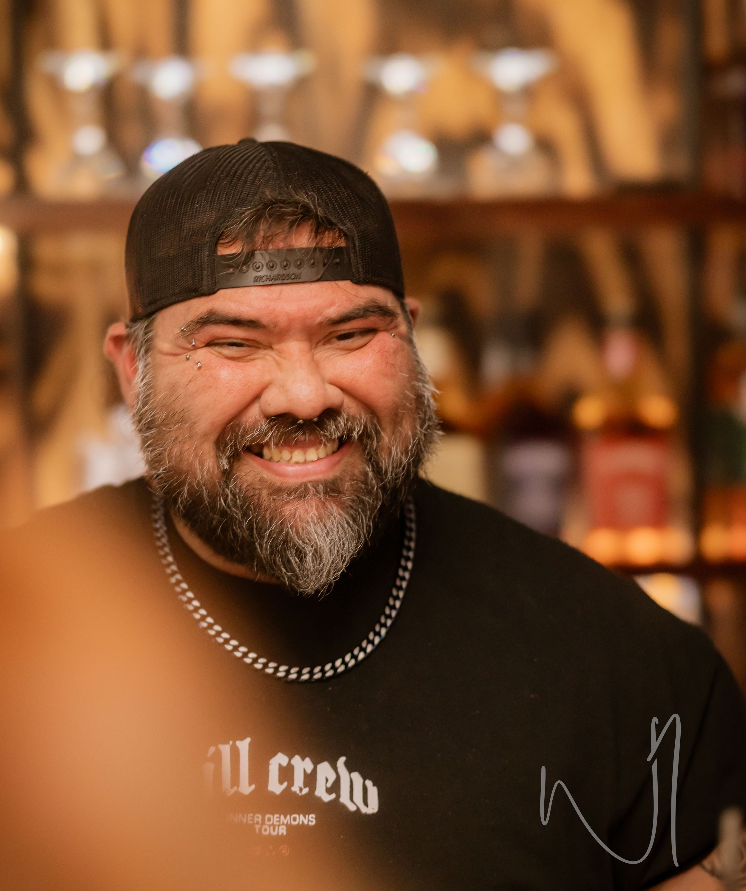 A bearded person wearing a black t-shirt with a chain necklace smiles warmly in a bar setting.