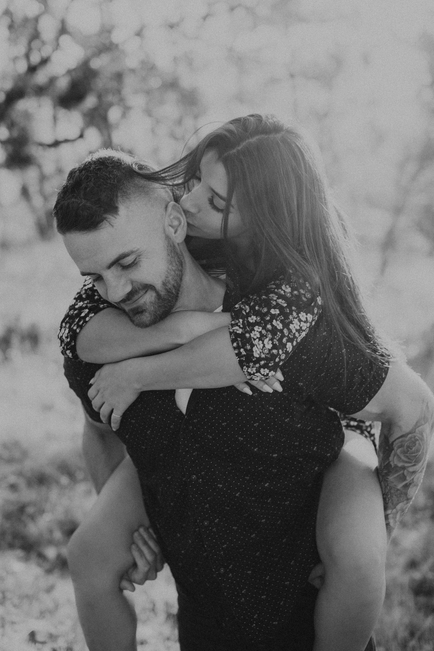 A couple shares an intimate black and white moment as she rides piggyback and kisses his cheek outdoors.