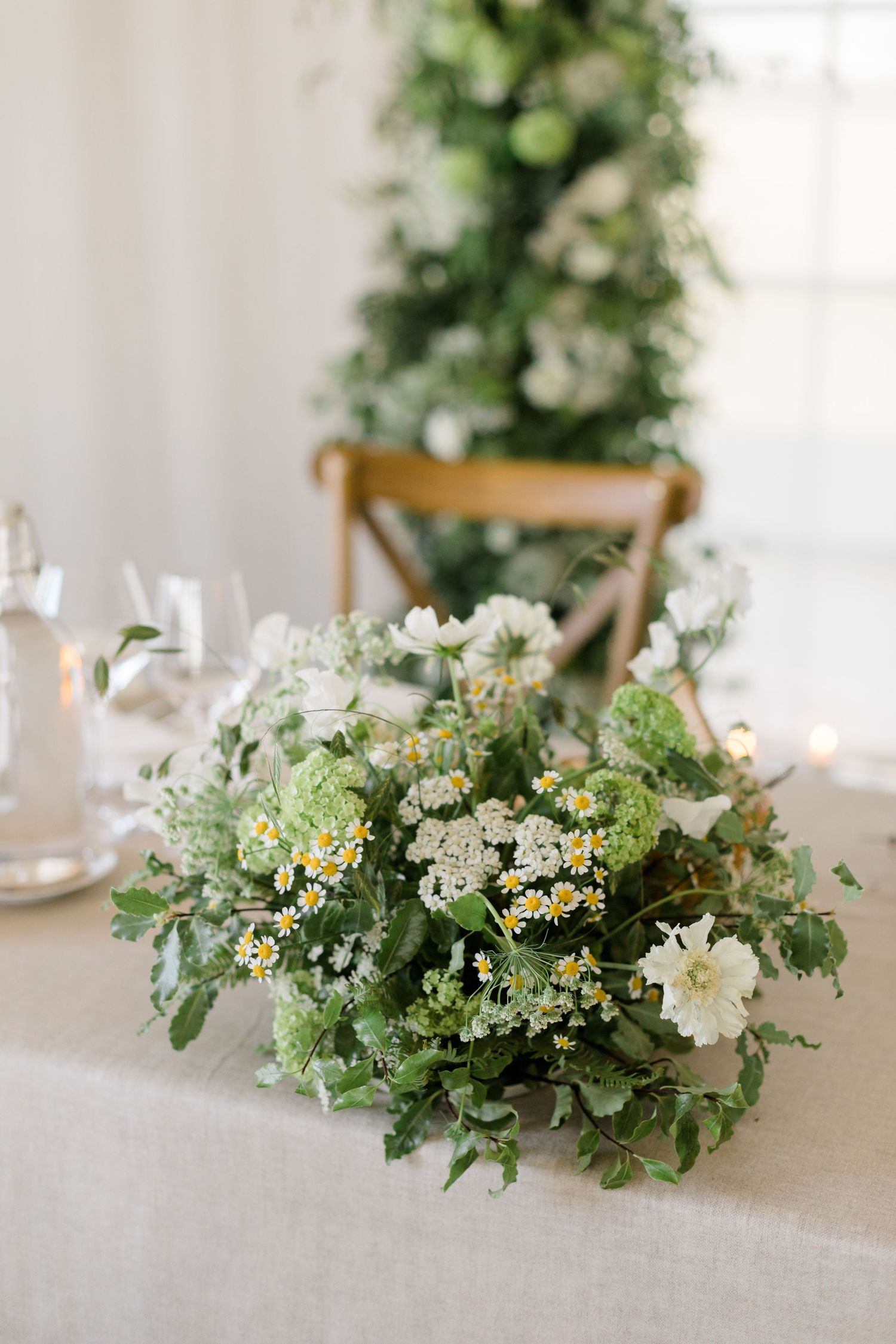 Delicate floral centerpiece with white daisies and greenery arranged on a white tablecloth in a bright dining setting.