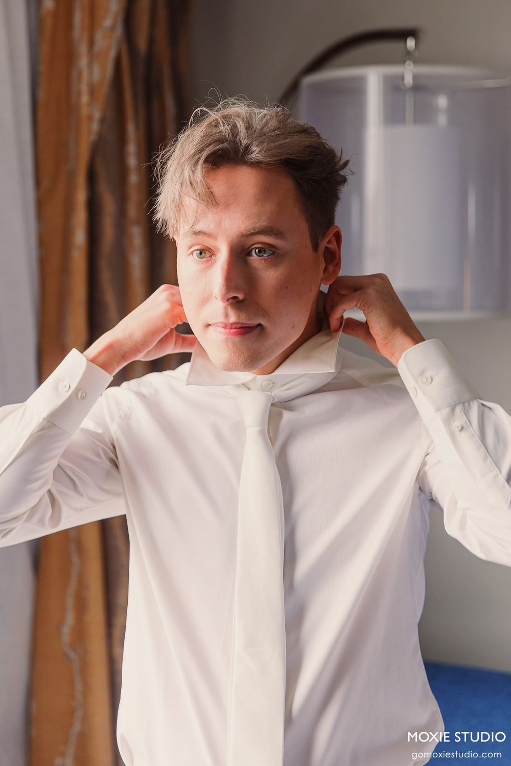 A person adjusts the collar of their crisp white dress shirt in a warm indoor setting.