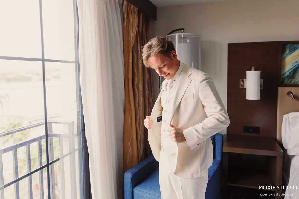 A person in a white suit jacket stands by a window in a hotel room, preparing for a formal event.