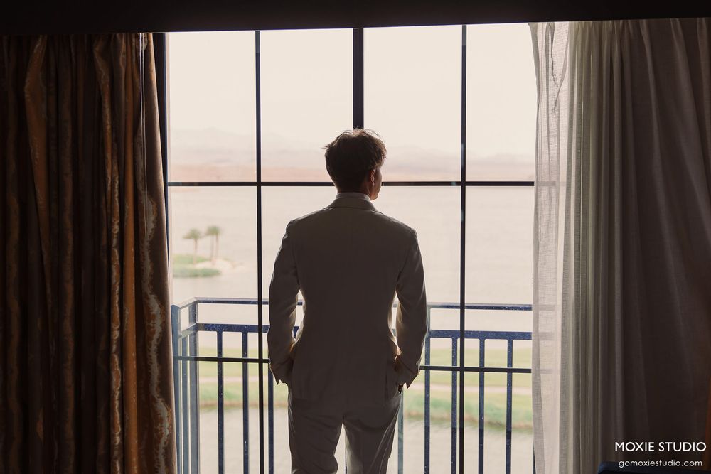 A silhouetted figure stands at a window with hands in pockets, gazing out at the view through paned glass.