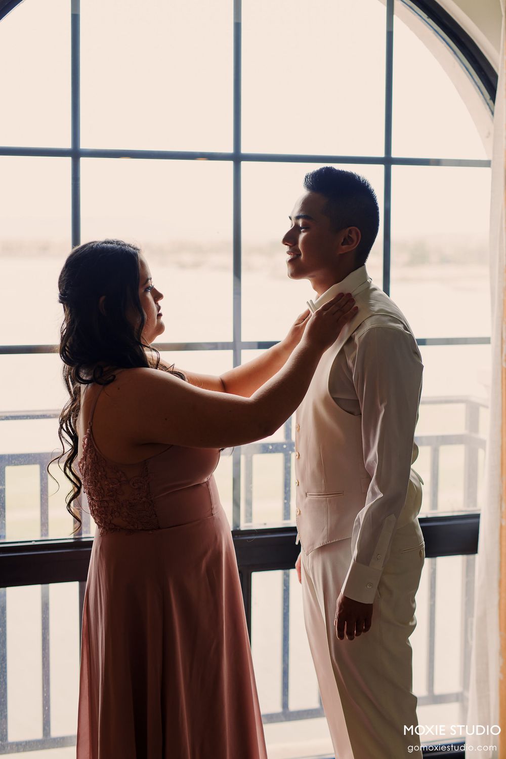 A couple shares an intimate moment by a large window as they prepare for a formal event.