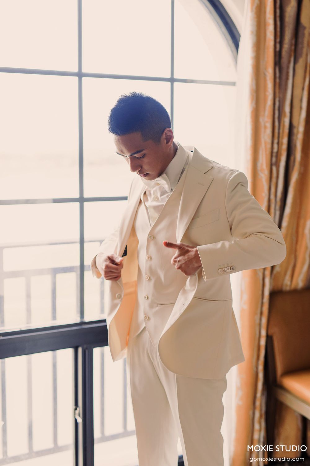 A person in a cream-colored suit jacket stands by a bright window in an elegant indoor setting.