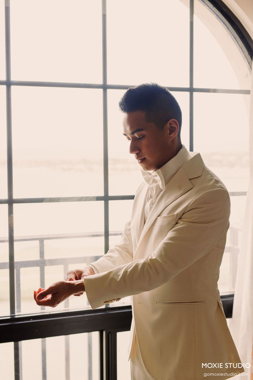 A person in a light beige suit jacket stands thoughtfully by a large window with geometric panes.