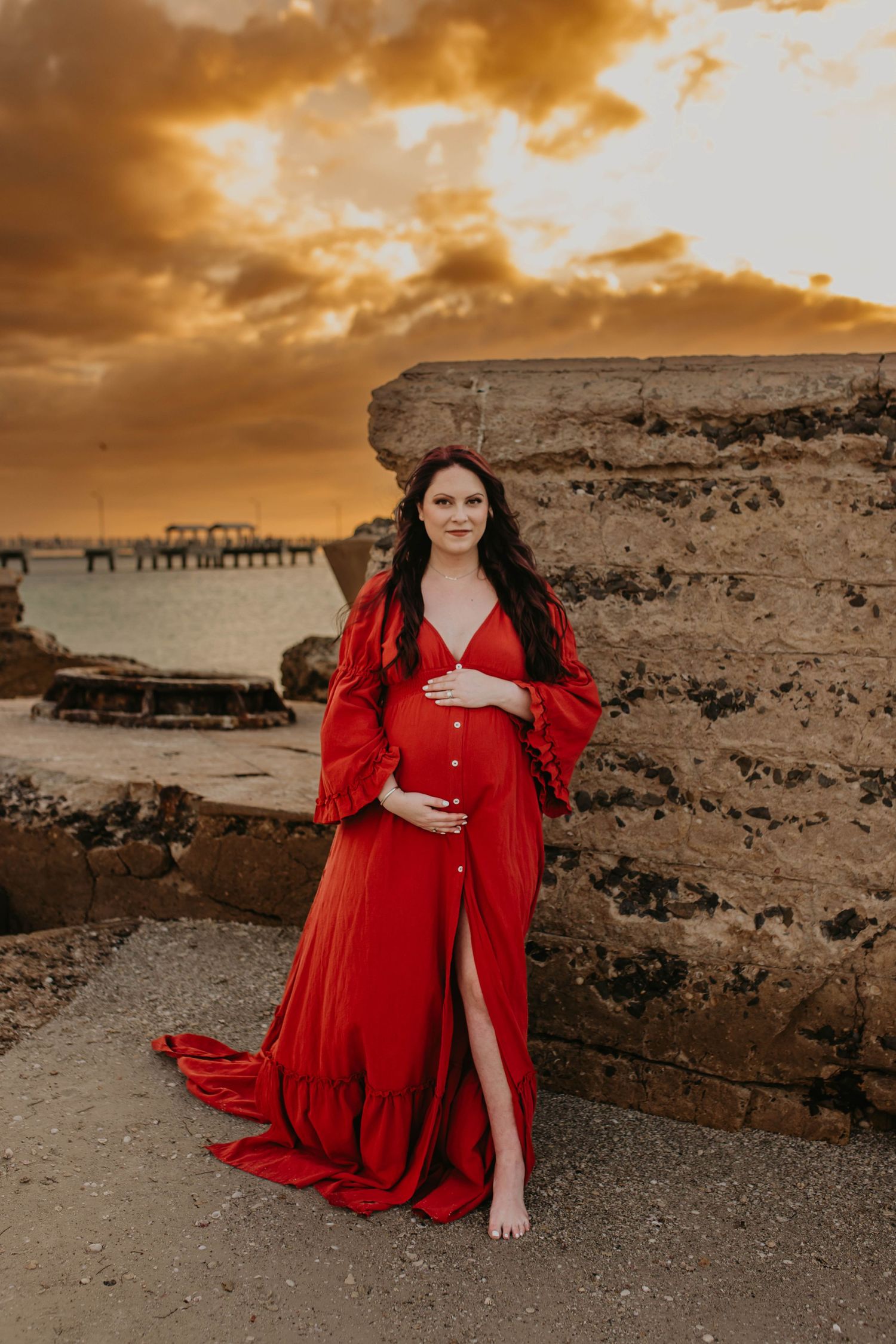 Maternity photo of woman in flowing red dress against dramatic coastal sunset backdrop.