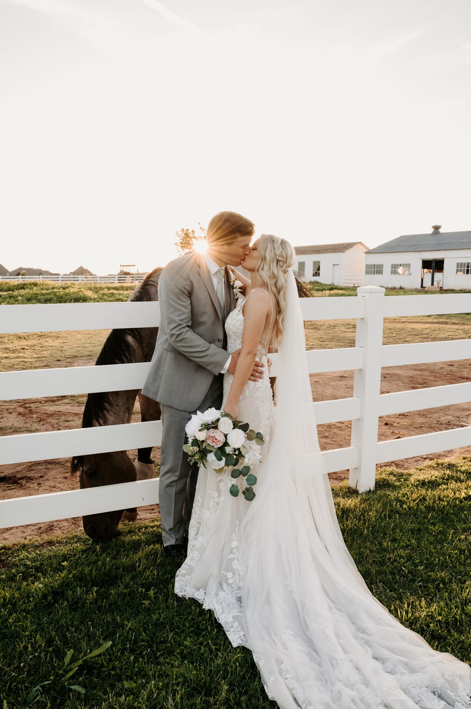 Wedding couple embraces by white fence during sunset at rustic farm venue. Oklahoma Wedding Photographer