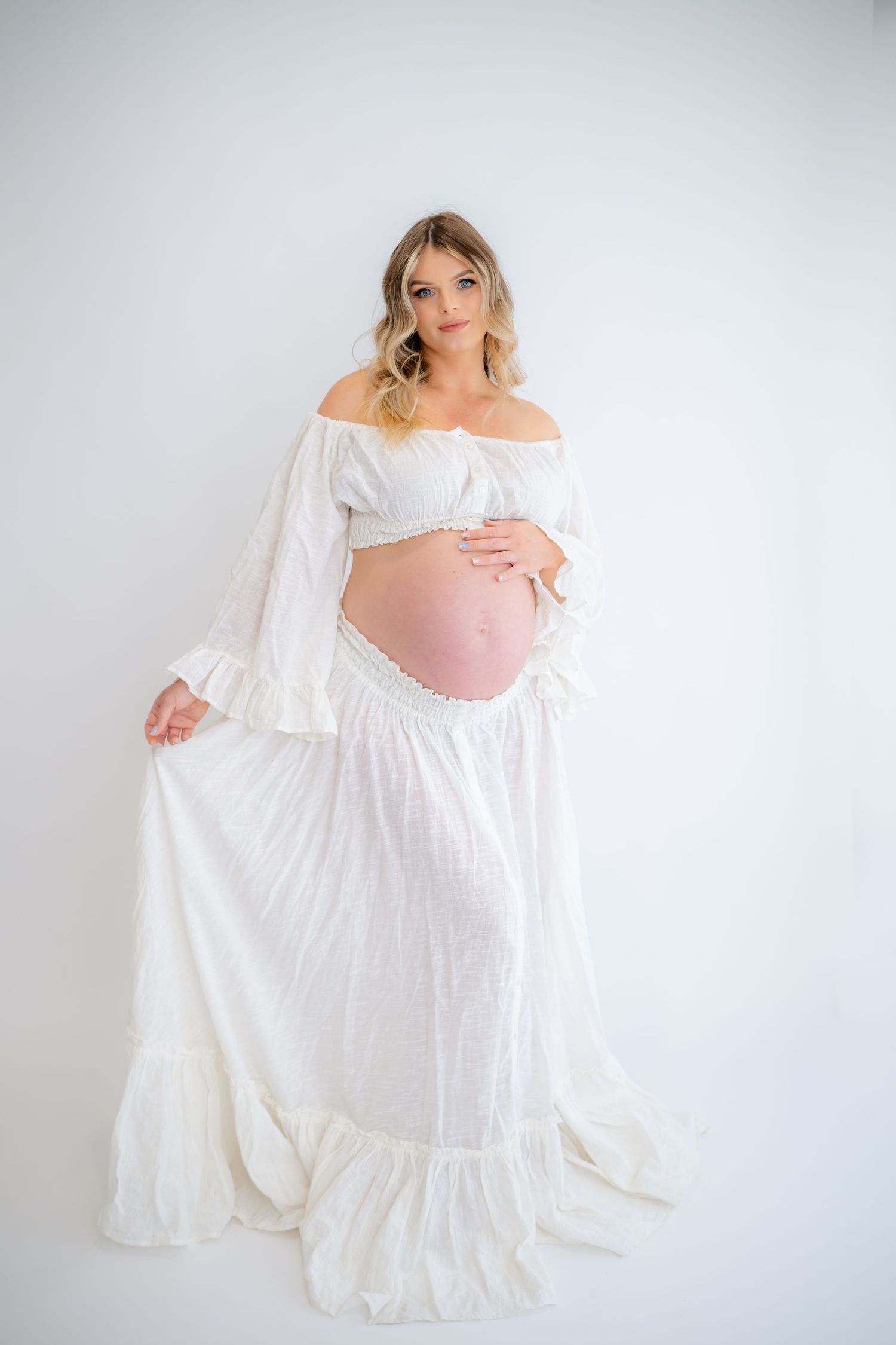 Maternity portrait in flowing white off-shoulder dress against light gray backdrop.
