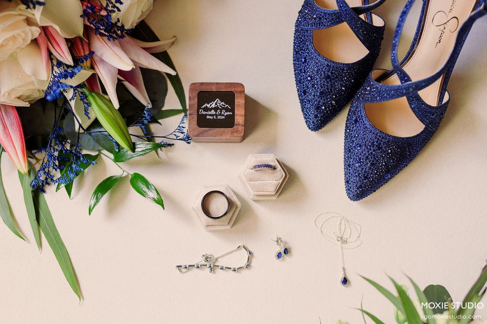 Wedding details including blue sparkly heels, floral arrangements, rings, and perfume bottle arranged on beige background.