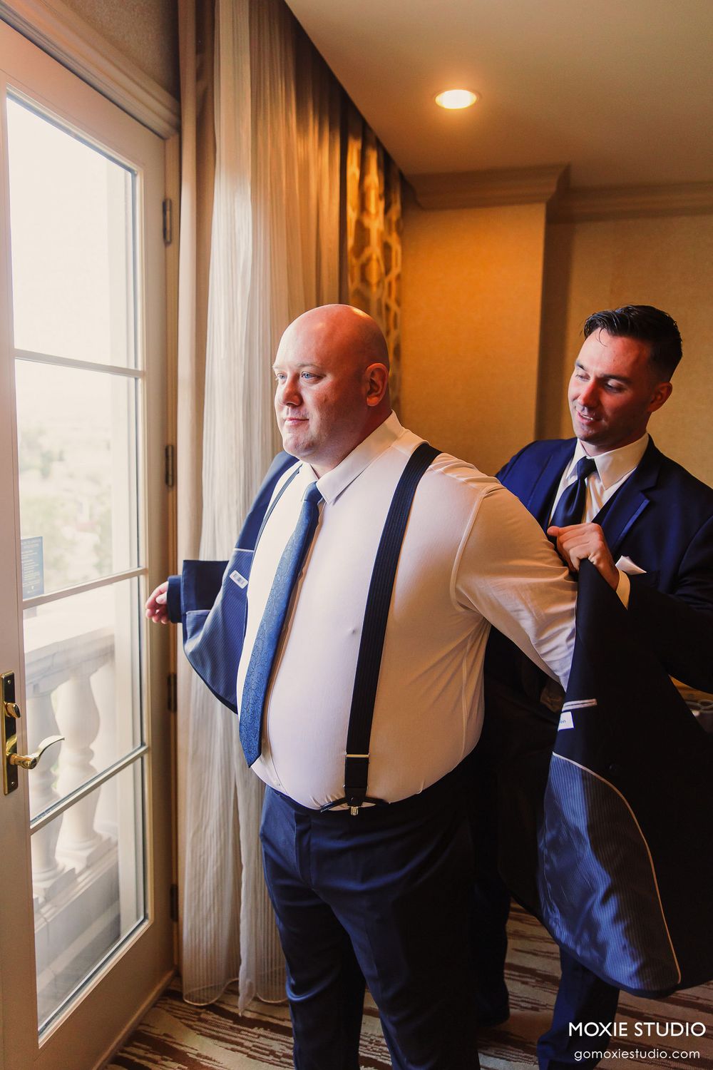 Groomsmen in navy suits and white shirts getting ready by a window in a warm-lit room before a wedding ceremony.