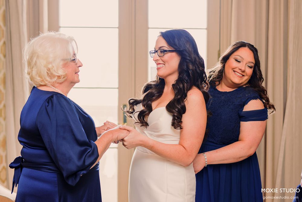 Three women share an emotional moment during wedding preparations in elegant navy and white dresses.