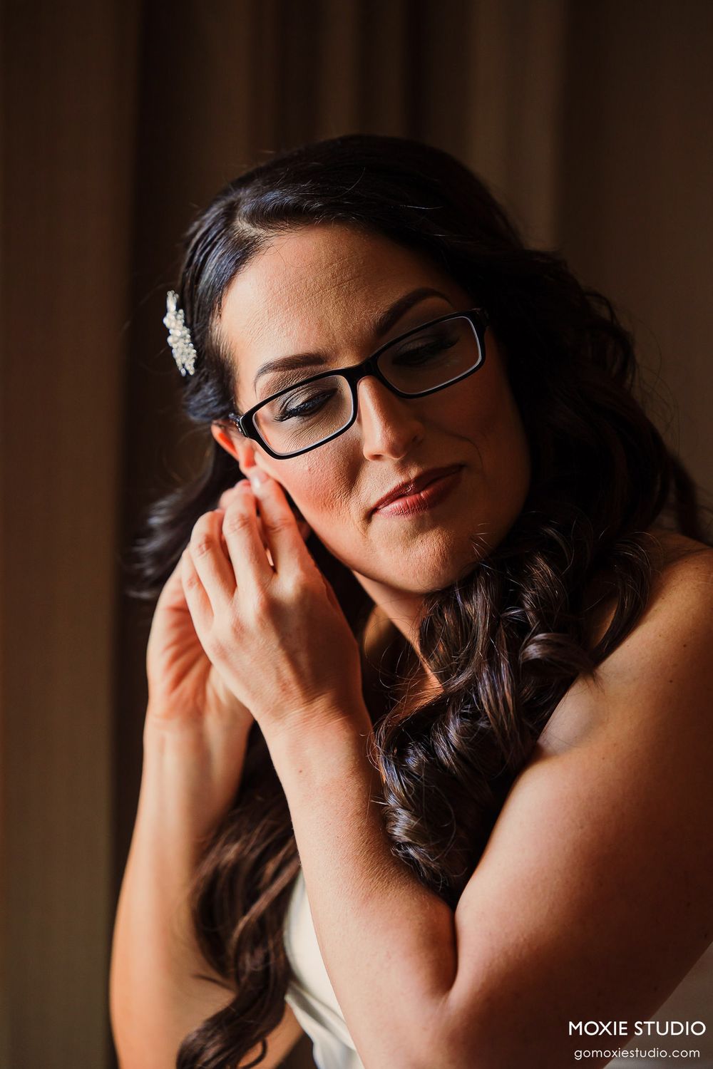 A contemplative bride in glasses and white gown touches her face in a gentle pose.