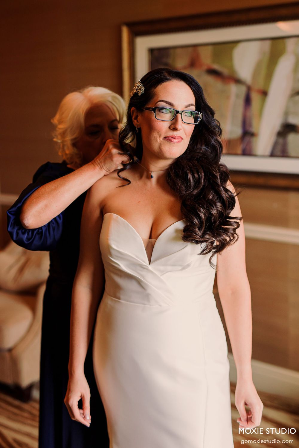 A bride in a white strapless gown with dark wavy hair and glasses gets ready for her wedding day.