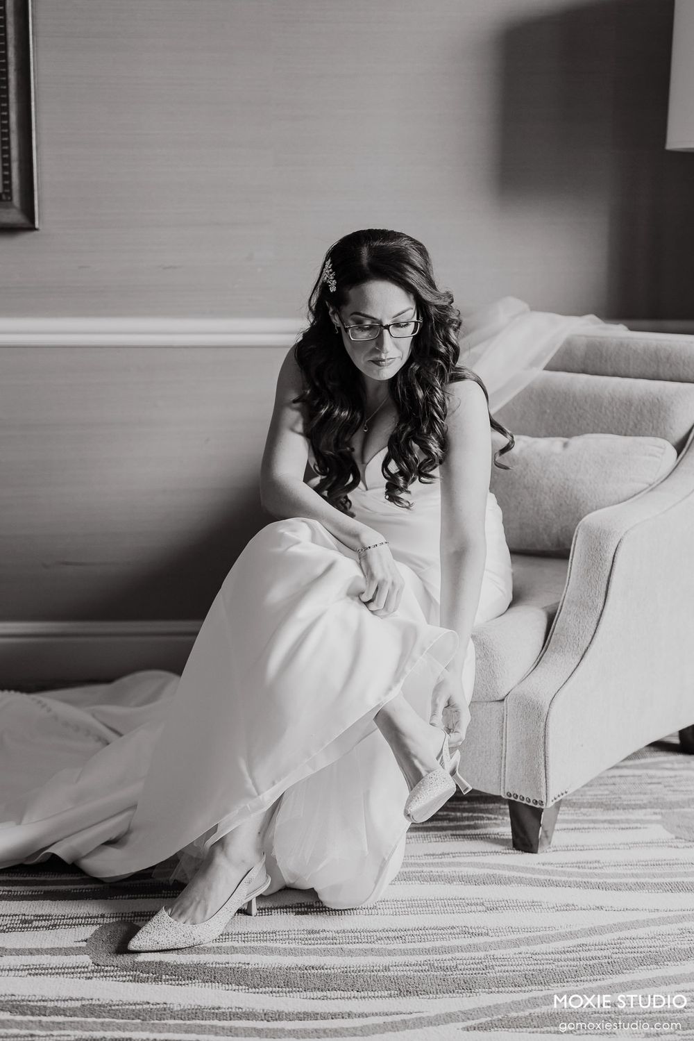 A bride in glasses and white gown sits elegantly on a light colored chair in black and white photography.
